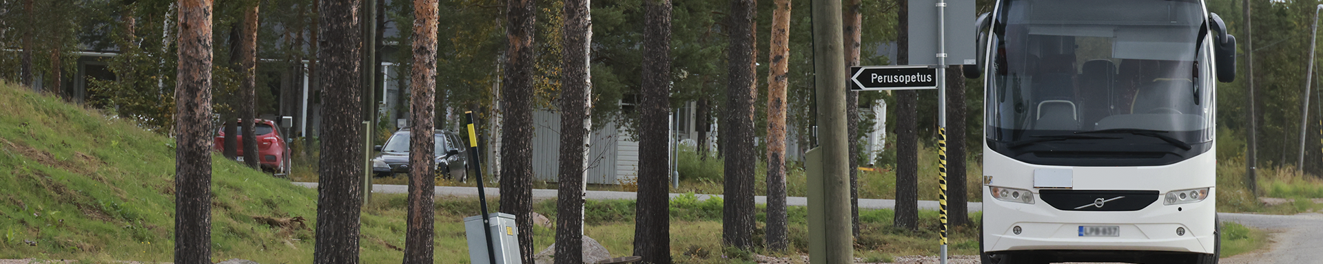 Valkoinen linja-auto on pysäköity tien varteen metsäisessä ympäristössä, jossa kasvaa korkeita puita. Bussin vieressä on kyltti, jossa lukee 'Perusopetus' ja osoittaa vasemmalle. Taustalla näkyy rakennuksia ja pysäköityjä autoja puiden lomassa, mikä viittaa maaseutu- tai lähiympäristöön koulun läheisyydessä.