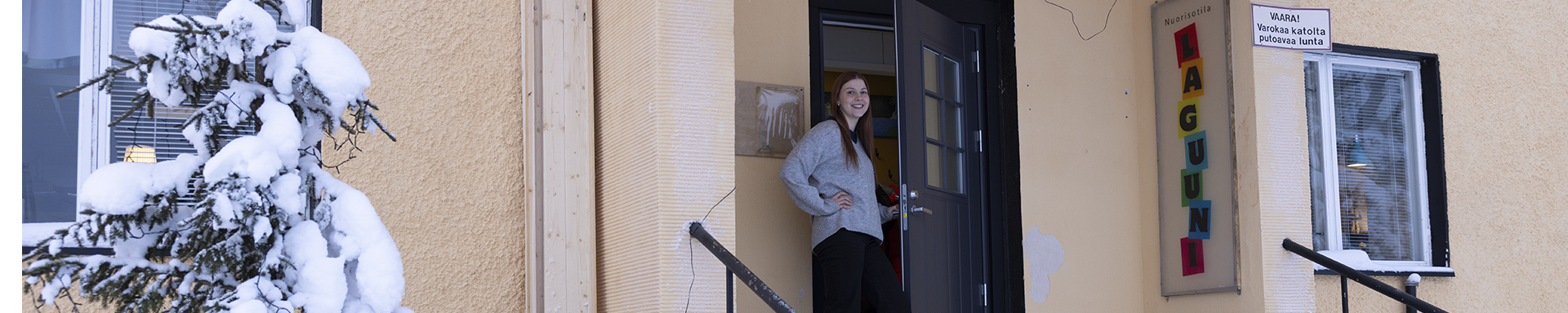 Henkilö seisoo rakennuksen sisäänkäynnin portailla talvisessa ympäristössä. Seinällä näkyy värikäs ‘Nuorisotila Laguuni’ -kyltti sekä varoitusmerkki katolta putoavasta lumesta. Portaiden vieressä on luminen kuusi ja taustalla rakennuksen ikkunoita.