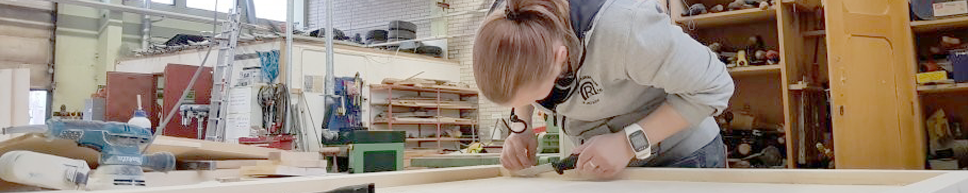 Henkilö työskentelee puuverstaassa, kumartuneena suuren puukappaleen ääreen ja käyttää työkalua mittaamiseen tai merkkaamiseen. Verstaassa on erilaisia työkaluja, tarvikkeita ja hyllyjä sekä tikkaat, mikä kuvaa puutyöskentelyn ympäristöä.