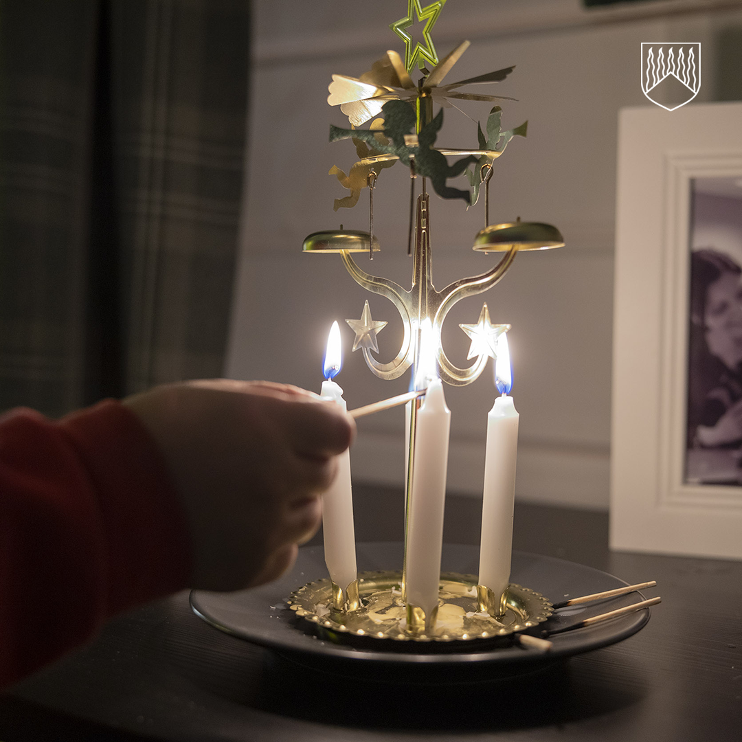 Käsi sytyttää valkoista kynttilää messinkisessä kynttelikössä, jossa on koristeellisia tähtiä ja pieniä pyöriviä metallisia hahmoja. Kynttelikkö seisoo mustalla lautasella, ja sen ympärillä on tulitikkuja. Taustalla näkyy kehystetty valokuva ja verhot. Kuvan oikeassa yläkulmassa on vaakunan kaltainen logo, jossa on pystysuuntaisia viivoja.