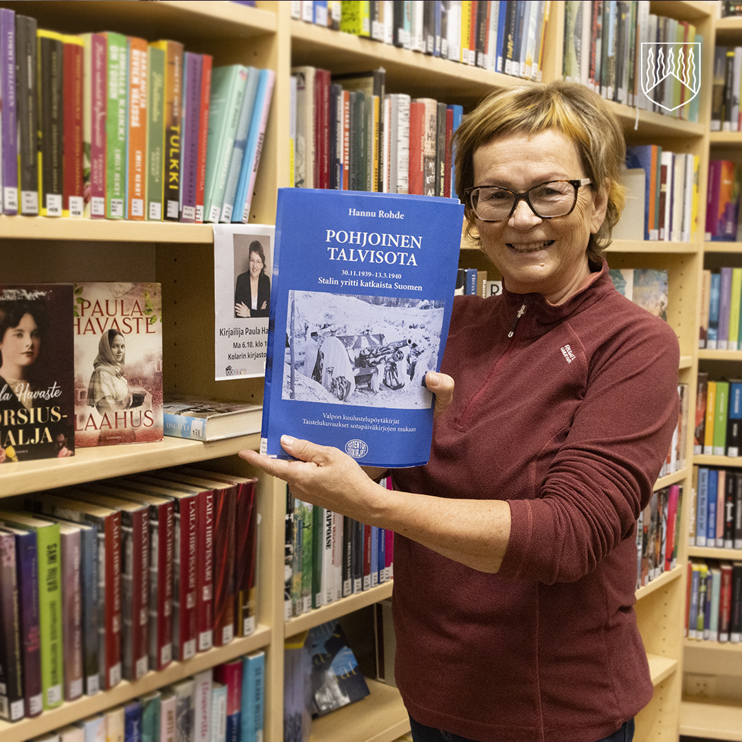 Sirpa Tolonen seisoo kirjastossa kirjahyllyn edessä ja pitelee sinikantista kirjaa 'Pohjoinen Talvisota' (Hannu Rautio). Kirjan kannessa on kuva talvivaatteisiin ja sotavarusteisiin pukeutuneista ihmisistä. Taustalla kirjahyllyssä näkyy kirjoja, muun muassa Paula Havasteen teokset 'Morsiusmalja' ja 'Laahus', sekä kyltti, jossa kerrotaan Paula Havasteen vierailusta kirjastossa.