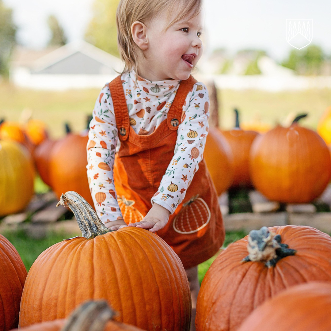 Lapsi seisoo kurpitsapellolla ja koskettaa suurta kurpitsaa. Hänellä on yllään oranssi kurpitsakuviollinen haalari ja syksyteemainen pitkähihainen paita. Taustalla näkyy lisää kurpitsoja ja ulkoilma-alue.