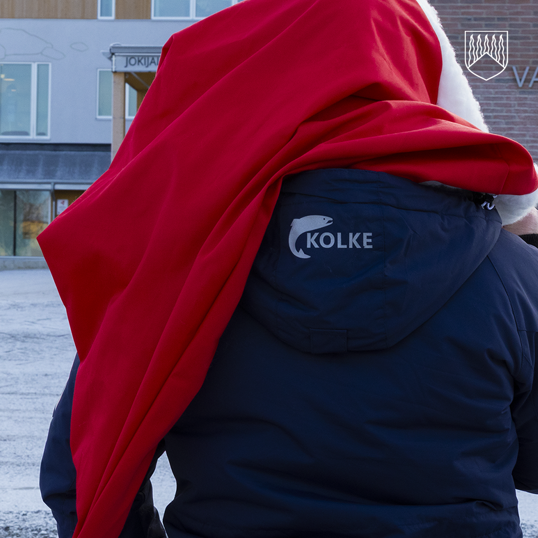 Henkilö seisoo ulkona talvisessa ympäristössä selkä kameraan päin. Henkilöllä on tummansininen takki, jonka selässä lukee “KOLKE” ja on kalan siluetti. Henkilön hartioilla on suuri punainen kangas, joka muistuttaa joulupukin viittaa, ja sen reunassa näkyy valkoista karvareunusta. Taustalla on harmaa rakennus, jossa on ikkunoita ja kyltti.