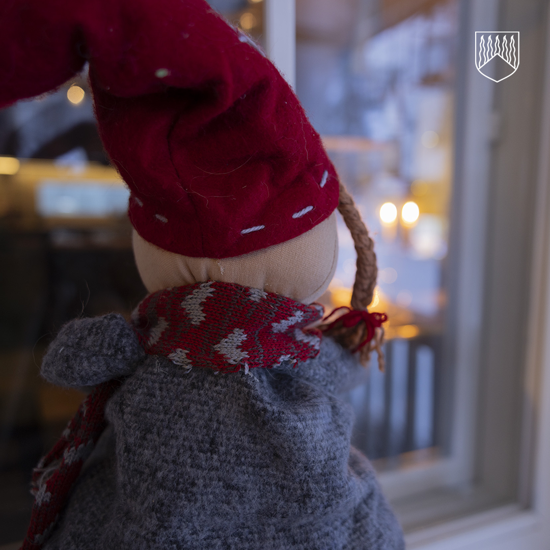 Pehmoinen tonttuhahmo kuvattuna takaapäin, pukeutuneena harmaaseen villatakkimaiselta näyttävään asuun ja punaiseen huiviin. Tontulla on suuri punainen lakki, jossa on valkoisia ompeleita, ja letitetty hiussuortuva. Hahmo katsoo ikkunasta ulos, taustalla näkyy kynttilänvaloa ja hämärä talvimaisema.