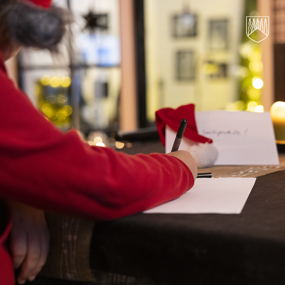 Henkilö pukeutuneena punaiseen asuun ja harmaaseen tonttulakkiin kirjoittaa kynällä valkoiselle paperille pöydän ääressä. Pöydällä on toinen paperi, jossa näkyy teksti ”Joulupukille!”, sekä punainen tonttulakki. Taustalla on ikkuna, josta näkyy valaistuja koristeita ja epätarkka joulukuusi, joka luo tunnelmallisen, jouluaiheisen miljöön.