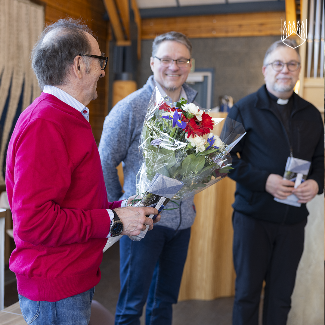 Kolme henkilöä seisoo sisätilassa. Etualalla oleva henkilö pitää käsissään kukkakimppua, ja kaksi muuta henkilöä seisoo taustalla puupintaisen seinän edessä.