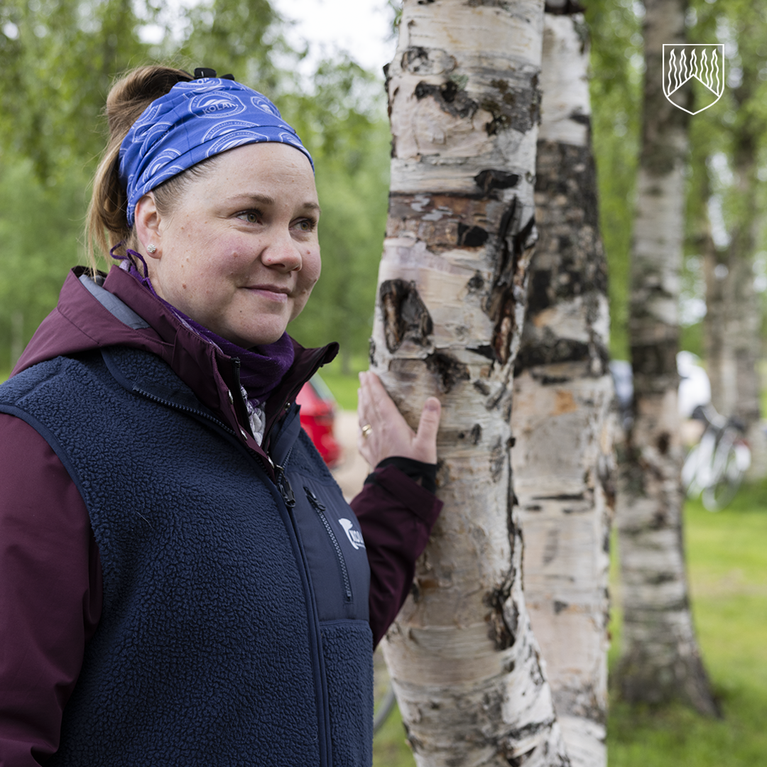 Kehitysjohtaja Nina-Maria Möykkynen seisoo ulkona koivujen keskellä ja pitää kättään puun rungolla. Henkilöllä on päässään sininen huivi sekä päällään ulkoiluun sopiva takki ja liivi. Taustalla näkyy vihreä ympäristö ja osittain parkkeerattu ajoneuvo.