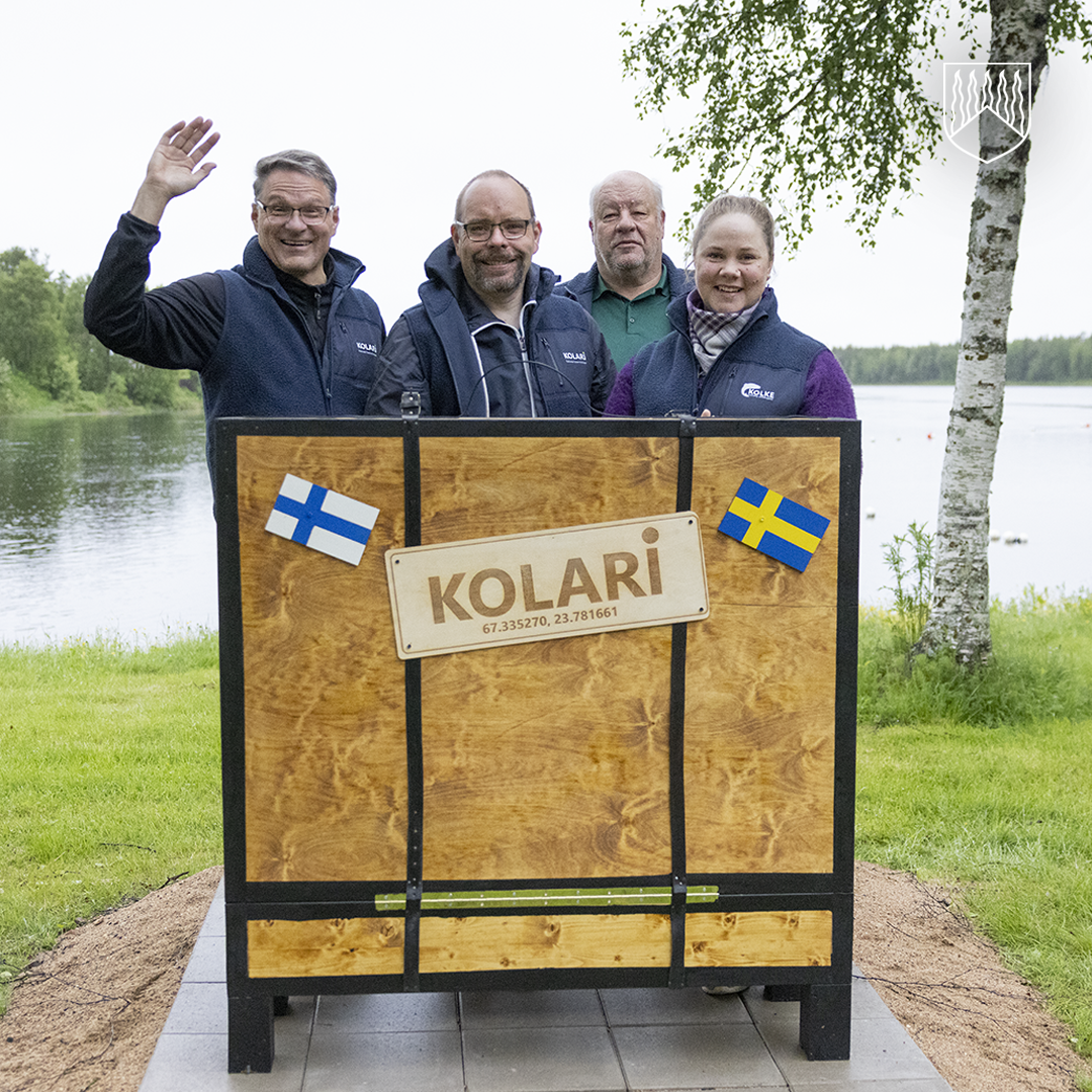 Kolarin kunnan johtoryhmän jäsenet Kolarinsaaren matkalaukkua esittävällä kuvauspisteellä joen rannalla. Kuvauspisteessä on Suomen ja Ruotsin liput sekä KOLARI-kyltti.