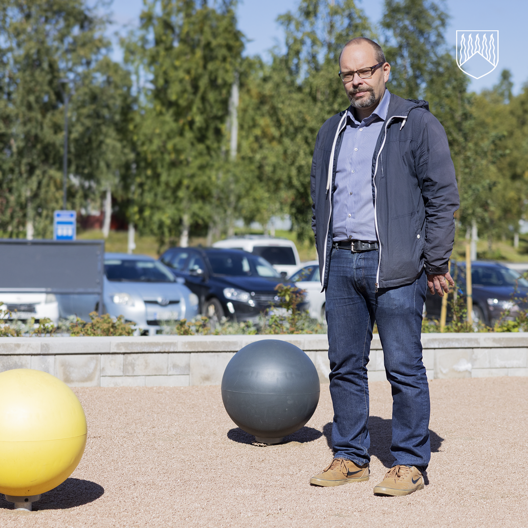 Ulkotiloissa sorapintaisella alueella seisova henkilö. Etualalla näkyy kaksi suurta pallomaista leikkipuisto- tai liikunta-alueen elementtiä, taustalla puita ja pysäköityjä autoja.