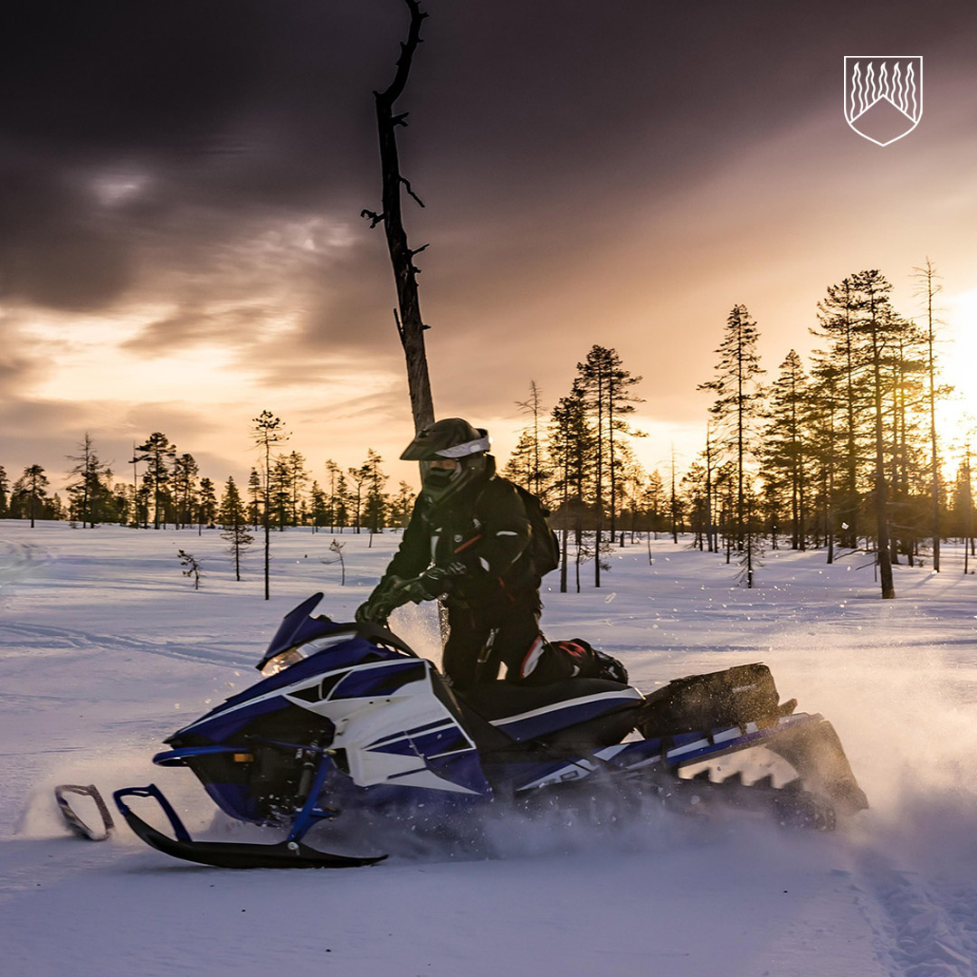 Moottorikelkkailija ajaa sinivalkoisella kelkalla lumisella suomaastolla auringon laskiessa. Taustalla näkyy harvakseltaan puita ja oranssinsävyinen taivas, jossa tummat pilvet luovat dramaattisen tunnelman.