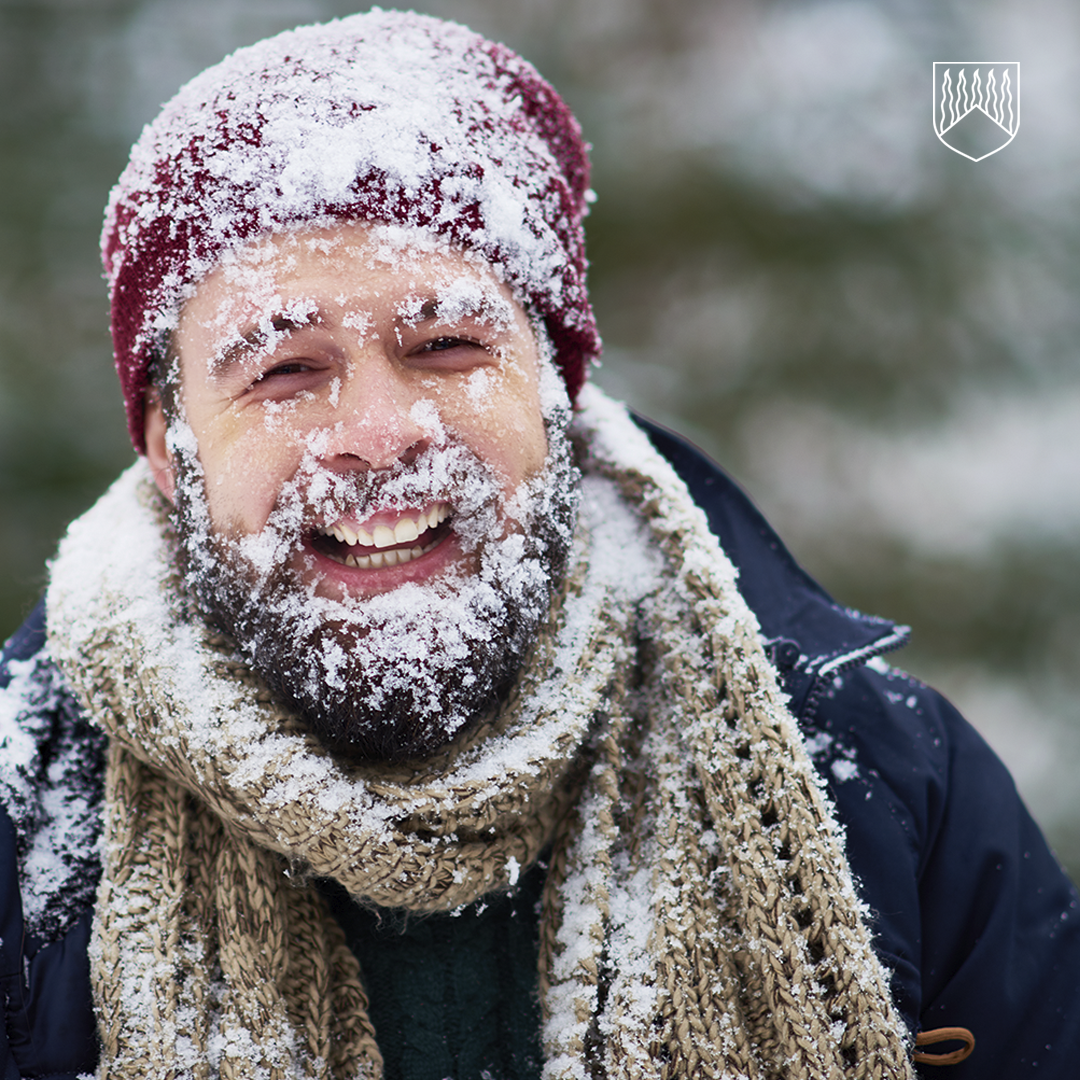 Henkilö pukeutuneena talvivaatteisiin, päässään punainen pipo ja kaulallaan paksu, vaaleanruskea neulehuivi. Vaatteissa ja pipossa on lumikerroksia, mikä viittaa lumisateeseen tai ulkoiluun talvisessa ympäristössä. Taustalla näkyy epätarkka, luonnon sävyinen maisema.