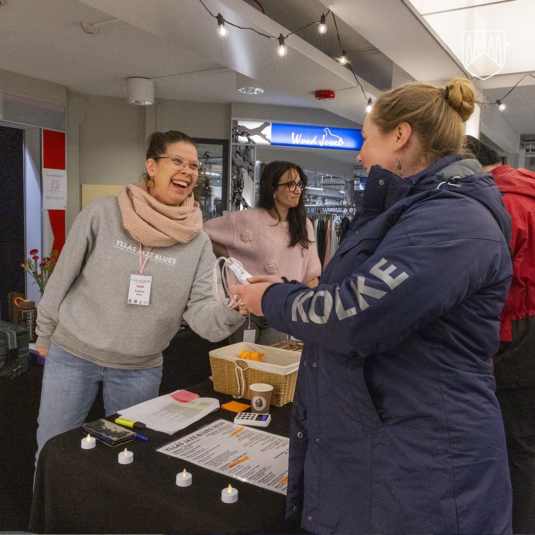 Tapahtuman infopiste, jossa henkilö ojentaa ranneketta toiselle osallistujalle. Pöydällä on listoja, kyniä, teevaloja ja kori tarvikkeille. Taustalla näkyy myymälä ja tapahtumavalaistus.